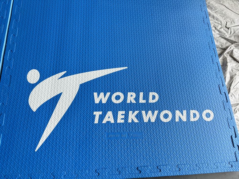 Surface detail of an embossed blue puzzle mat printed with the World Taekwondo mark, interlocking edge tabs visible along the sides